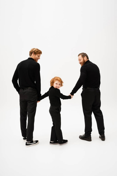 full length of cheerful redhead boy looking at camera while holding hands with dad and grandpa in black clothes on light grey background - Photo, image