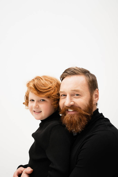 happy bearded man and redhead grandson wearing black sweaters and looking at camera isolated on grey - Photo, Image