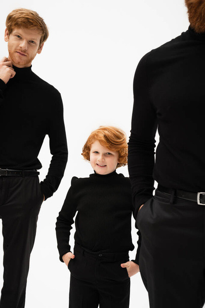 cheerful kid in black clothes looking at camera while posing with hands in pockets near redhead father and grandpa isolated on grey - Zdjęcie, obraz