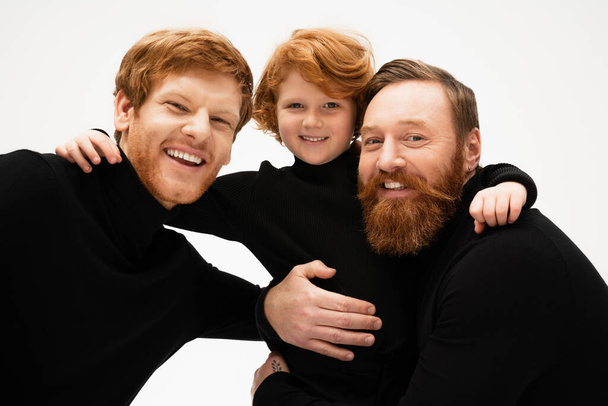 joyful redhead boy with father and bearded granddad embracing while looking at camera isolated on grey - Photo, image