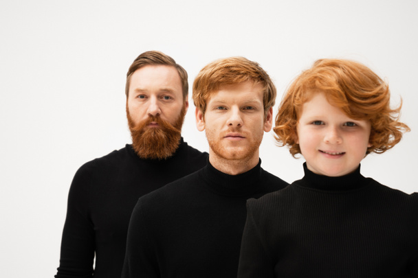 happy red haired boy smiling at camera near father and bearded granddad in black sweaters isolated on grey - Photo, image