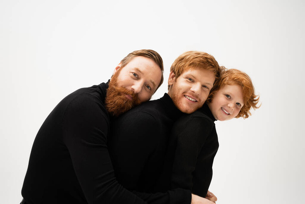 cheerful redhead men and kid in black sweaters leaning on each other and smiling at camera isolated on grey - Zdjęcie, obraz