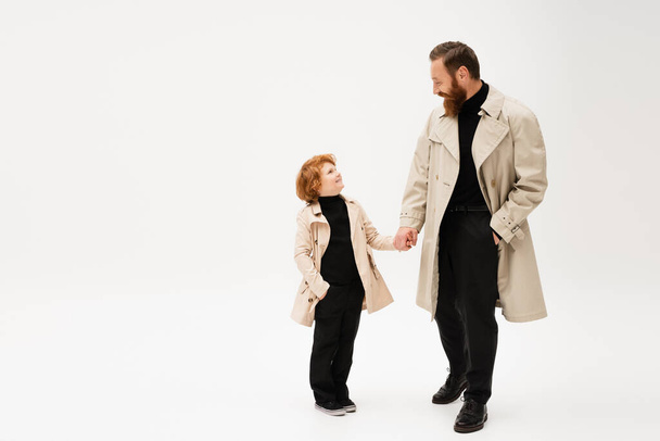 happy redhead kid and bearded man in trench coats holding hands and looking at each other on light grey background - Photo, image