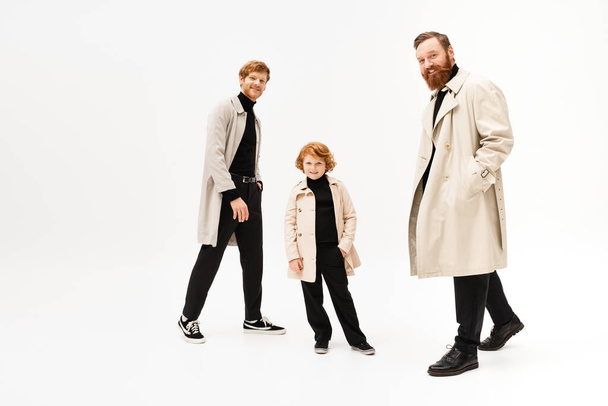 full length of happy redhead men and kid in trench coats looking at camera while posing on light grey background - Fotó, kép