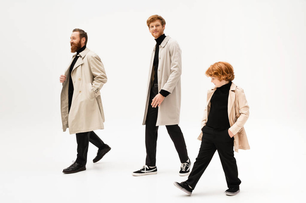 young man in trench coat smiling at camera while walking near bearded father and redhead son on light grey background - Foto, Imagem