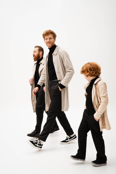 full length of trendy bearded men and redhead boy in trench coats walking on light grey background - Photo, image