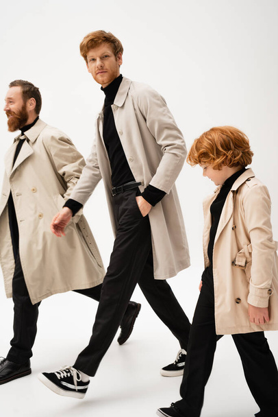 молодий чоловік в тренч пальто дивиться на камеру під час ходьби з бородатим батьком і рудим сином на світло-сірому фоні
 - Фото, зображення