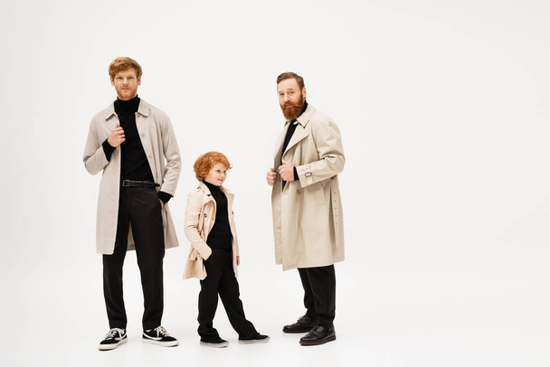 full length of stylish redhead men and boy in trench coats posing on light grey background - Foto, imagen