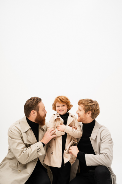 happy redhead kid in trench coat smiling near father and bearded grandpa hugging him isolated on grey - Foto, Imagen