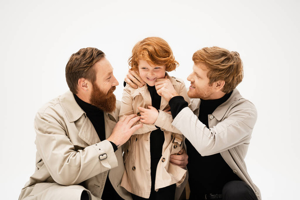 happy bearded men in trench coats embracing cheerful redhead boy isolated on grey - Photo, image