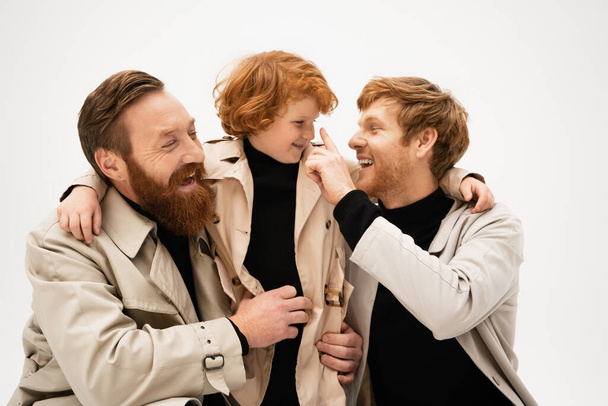 cheerful redhead men in trench coats embracing and having fun with smiling boy isolated on grey - Foto, Bild