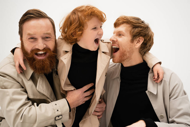 χαρούμενους γενειοφόρους άνδρες και κόκκινα μαλλιά αγόρι σε καμπαρντίνες γέλιο απομονωμένο σε γκρι - Φωτογραφία, εικόνα