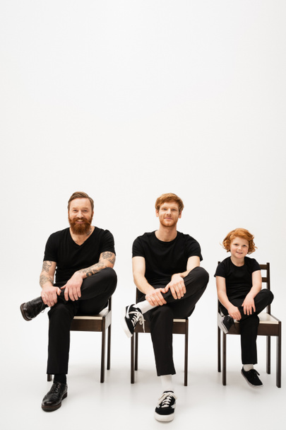 cheerful red haired boy with bearded grandfather and dad sitting on chairs and smiling at camera on grey background - 写真・画像