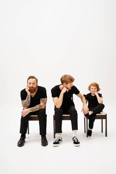 bearded tattooed man smiling at camera near redhead son and grandson sitting on chairs on grey background - Fotografie, Obrázek