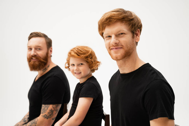 smiling man with redhead son and bearded tattooed father sitting and looking at camera isolated on grey - Foto, Bild