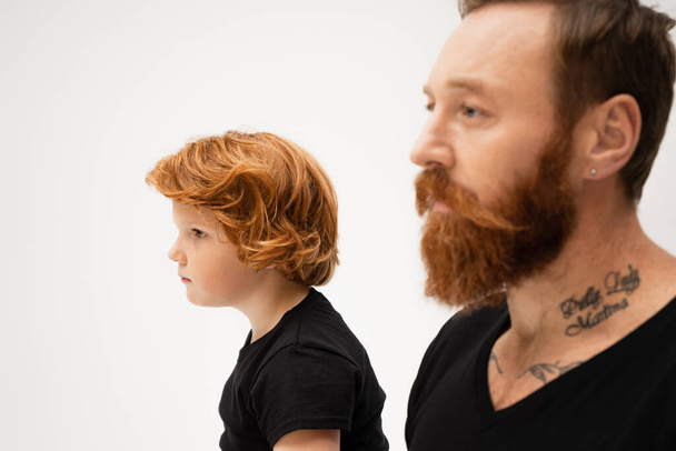 side view of redhead boy in near bearded and tattooed grandpa on blurred foreground isolated on grey - Zdjęcie, obraz