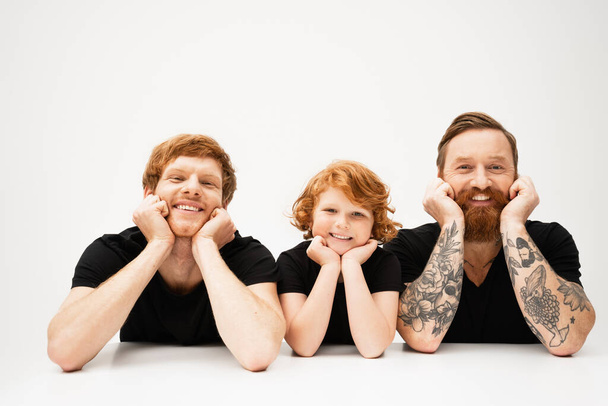 happy boy with redhead father and bearded tattooed grandpa lying and smiling at camera on light grey background - Foto, imagen