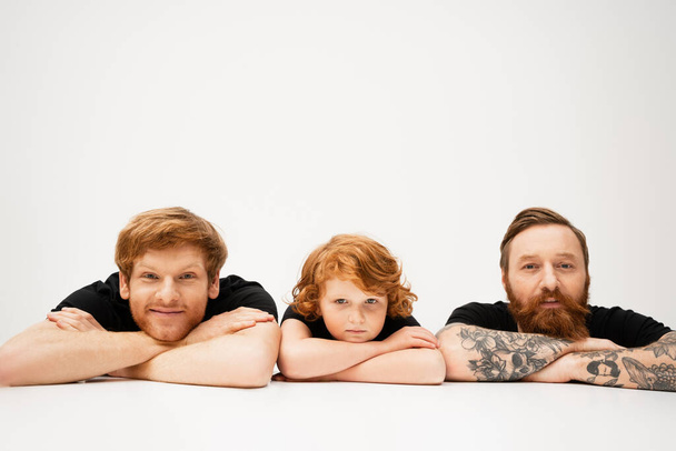 cheerful man with redhead son and bearded tattooed father lying with crossed arms on light grey background - Photo, image