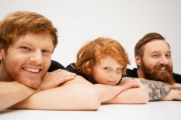 joyful man with redhead son and bearded tattooed dad lying with crossed arms and smiling at camera on light grey background - Photo, image
