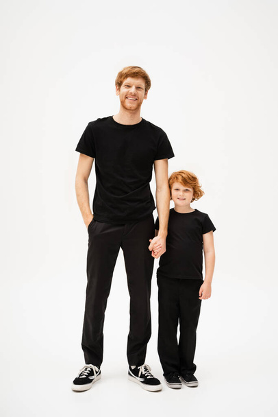 full length of cheerful red haired dad and son holding hands and looking at camera while standing on light grey background - Fotó, kép
