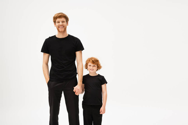 joyful redhead father and son in black t-shirts and trousers holding hands and smiling at camera isolated on grey - Foto, Imagen