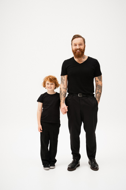 full length of bearded tattooed man with redhead grandson holding hands and smiling at camera on light grey background - Foto, Imagem