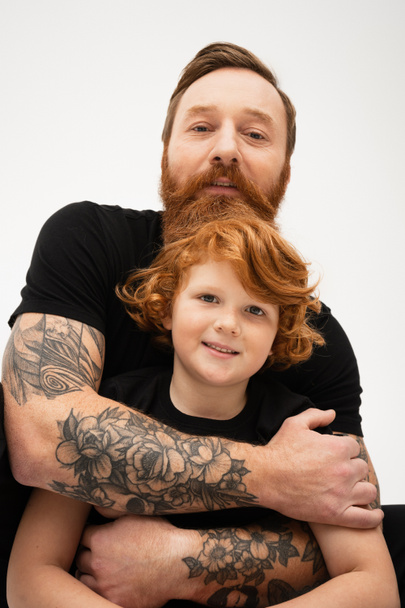 tattooed man in black t-shirt hugging red haired grandson smiling at camera isolated on grey - Photo, image