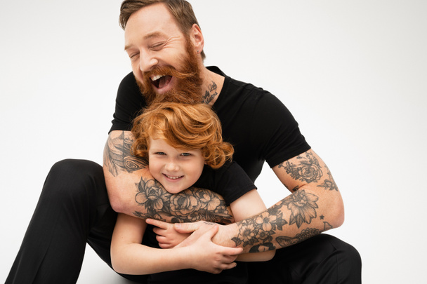 veselý tetovaný muž se zavřenýma očima objímající šťastný zrzka vnuk na šedém pozadí - Fotografie, Obrázek