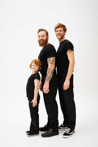 full length of bearded redhead men and kid in black t-shirts and trousers smiling at camera while standing on light grey background - Photo, Image