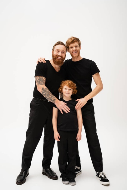 full length of joyful bearded men and redhead kid in black t-shirts and trousers posing on light grey background - Фото, зображення