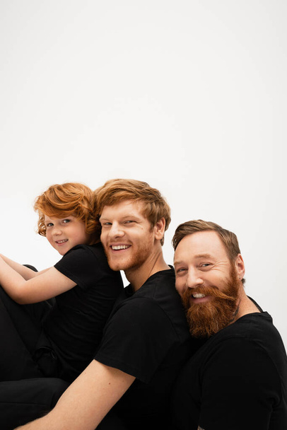 happy bearded man with redhead son and grandson in black t-shirts smiling at camera isolated on grey - Foto, Imagem