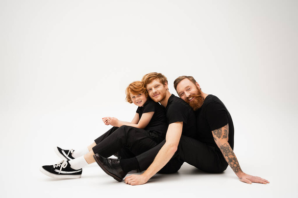 joyful redhead men and boy in black clothes looking at camera while sitting on light grey background - Foto, immagini