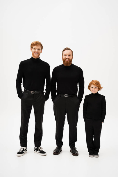 полная длина веселых бородатых мужчин с рыжими волосами ребенка в черной одежде, стоящих с руками в карманах на светло-сером фоне - Фото, изображение