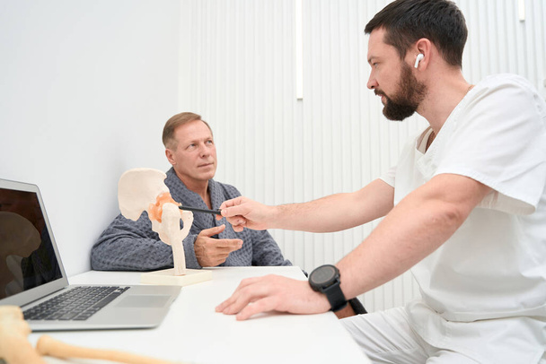 Professional orthopaedist pointing to a part of hip-bone replica on tabletop while talking to patient - Foto, Imagen