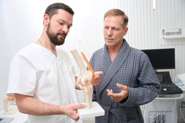 Qualified orthopaedist demonstrating a model of human knee joint to his patient - Fotó, kép