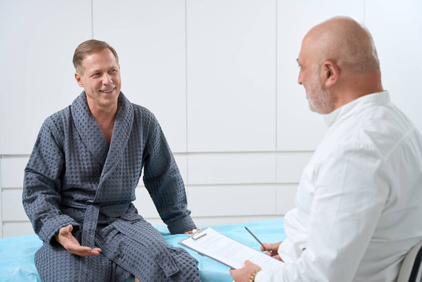 Hastane önlüklü bir adam tulumlu bir doktorun önünde oturuyor ve ameliyattan önce muayene oluyor. - Fotoğraf, Görsel