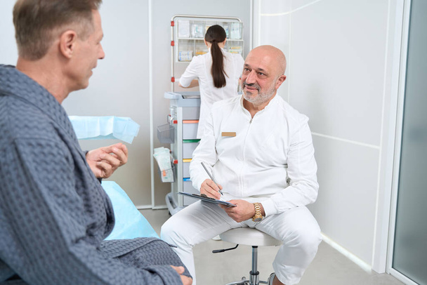 Внимательный врач общается с пациентом среднего возраста в клинике, они расположены в светлой комнате - Фото, изображение