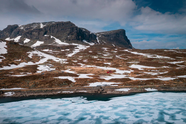 Snowy landscape of Hardangervidda with mountains and icy lake in Norway, red hut at the lakeside from above - Fotó, kép