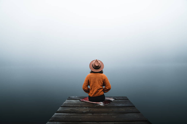 Back view of fashioned young woman sitting on wooden dock looking at view on a misty morning. Female hipster with brown hat relaxes on the edge of jetty admiring foggy lake. Wonderful nature getaway - Photo, image