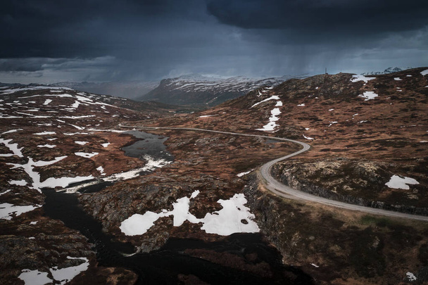 Lonesome road through the landscape of Hardangervidda National Park in Norway, snow fields and dark clouds in the sky, from above - Fotó, kép