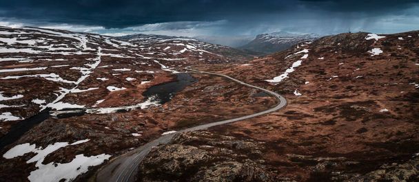 Lonesome road through the landscape of Hardangervidda National Park in Norway, snow fields and dark clouds in the sky, from above - Zdjęcie, obraz