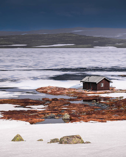 Hut at the waterfront of a frozen lake in the landscape of Hardangervidda National Park in Norway, snow and ice on the ground - Foto, Imagem