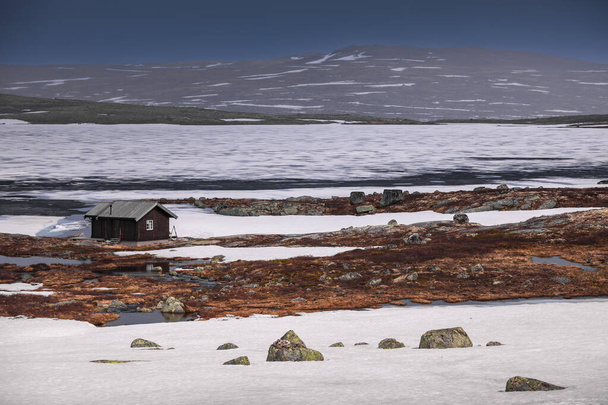 Hut at the waterfront of a frozen lake in the landscape of Hardangervidda National Park in Norway, snow and ice on the ground - Fotó, kép