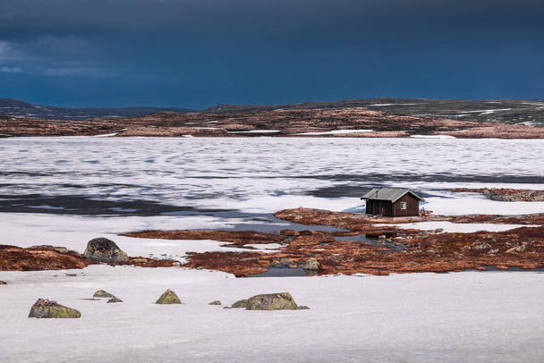 Hut at the waterfront of a frozen lake in the landscape of Hardangervidda National Park in Norway, snow and ice on the ground - Foto, imagen