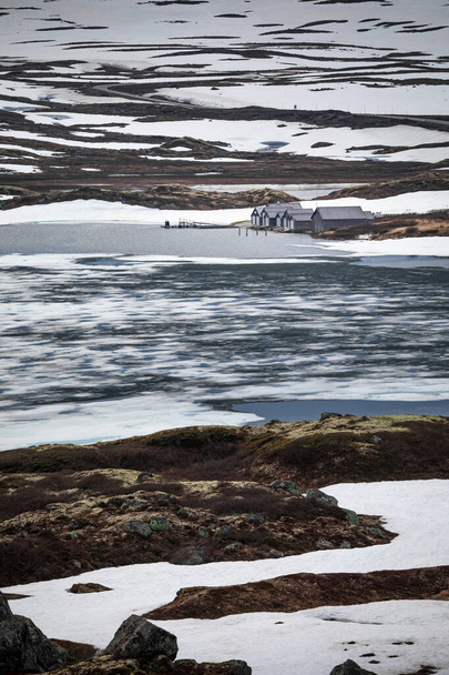 Cabins at the waterfront of a frozen lake in the landscape of Hardangervidda National Park in Norway, snow and ice on the ground - Fotó, kép