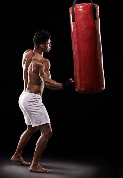 Strike while the iron is hot. Studio shot of kick boxer working out with a punching bag against a black background - Photo, image