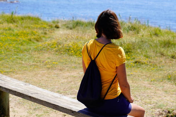 kobieta turysta siedzi na drewnianej ławce patrzy morze Atlantycki ocean na spacerze plaża lato wycieczka z plecakiem - Zdjęcie, obraz