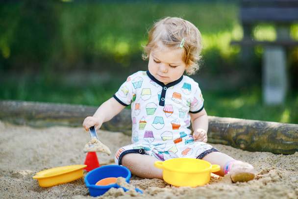 Linda niña jugando en la arena en el patio al aire libre. Hermoso bebé en ropa de verano que se divierte en el soleado día cálido de verano. Actividad al aire libre para niños pequeños - Foto, imagen