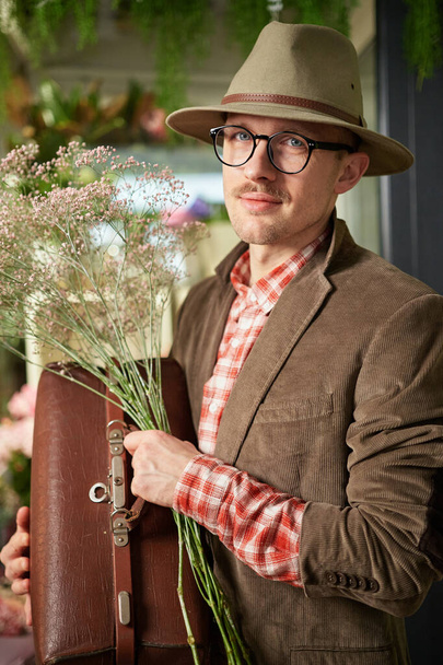 Koncept Valentýna. Veselý kavkazský mužský básník nebo spisovatel v brýlích a veteránských šatech s kloboukem, který zůstává s květinovou kyticí a dívá se do kamery v květinářství. Vysoce kvalitní vertikální obrázek - Fotografie, Obrázek