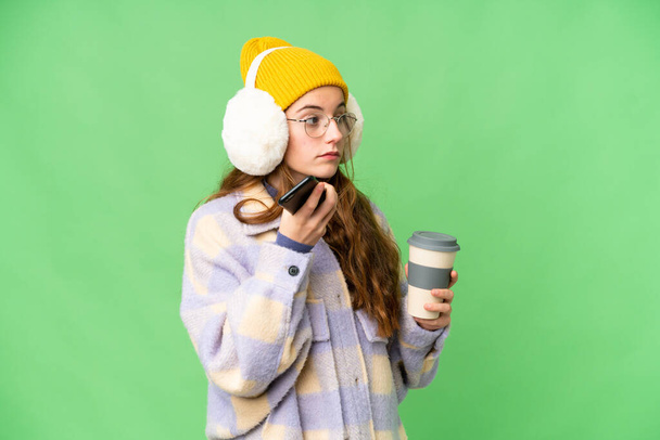 Teenager girl wearing winter muffs over isolated chroma key background holding coffee to take away and a mobile - Fotó, kép